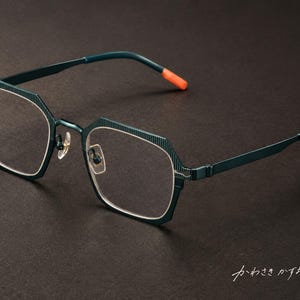 Puede incluir: Gafas de color verde azulado oscuro con montura octogonal que presenta una fina textura estriada vertical en el borde exterior. Las varillas son finas y del mismo color, con puntas de color naranja brillante. El puente y las plaquetas son plateados.