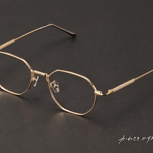May include: Gold-toned metal eyeglasses featuring a distinct octagonal frame shape, thin temples with a ribbed texture near the hinges, and clear lenses. The Japanese text "かわさきかずお" is visible in the bottom right corner of the frame.
