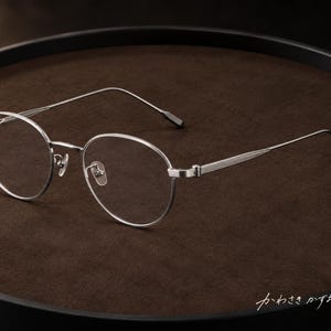 以下が含まれることがあります： 丸いフレームと細いテクスチャー加工が施されたテンプルを持つシルバーカラーの金属製メガネ。ブリッジには調整可能なノーズパッドがあり、テンプルの先端はダークカラーです。右下に日本語の「かわさきかずお」という文字が見えます。