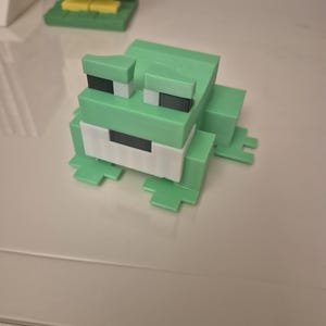 Op de afbeelding: Een blokkerig, gepixeld kikkerfiguur opgebouwd uit lichtgroene kunststof onderdelen. De kikker heeft een wit rechthoekig mondgedeelte, donkergrijze ogen en een gesegmenteerd lichaamsontwerp dat een digitale esthetiek met lage resolutie nabootst.