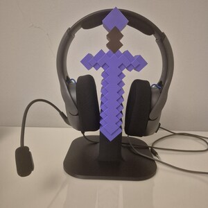 May include: A black gaming headset resting on a stand shaped like a pixelated purple sword. The sword features a dark brown handle and a square pommel. The headset includes a flexible boom microphone with a black foam cover and a wired connection.