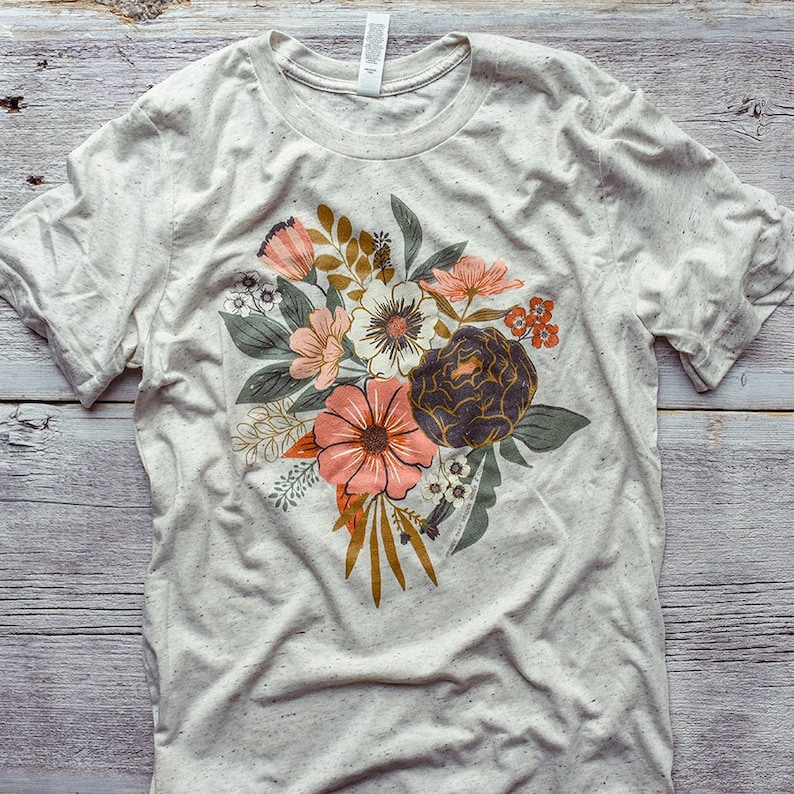 May include: A light gray t-shirt with a floral print featuring a bouquet of pink, white, and yellow flowers with green leaves and gold stems.