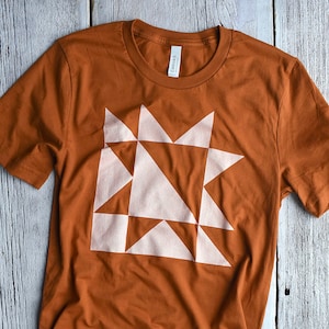 May include: A brown t-shirt featuring a white geometric design reminiscent of a quilt block. The design is composed of triangles and squares.