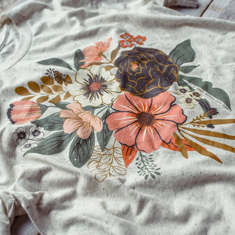 May include: A white t-shirt with a floral print featuring pink, white, and green flowers. The flowers are arranged in a bouquet shape and have a vintage style.