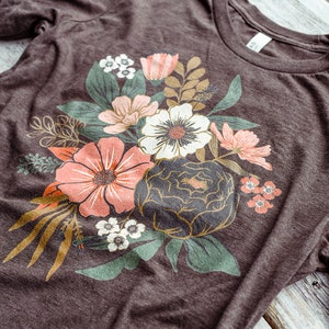 May include: A brown t-shirt with a floral print featuring a variety of flowers in shades of pink, white, and green. The flowers are arranged in a bouquet-like design.