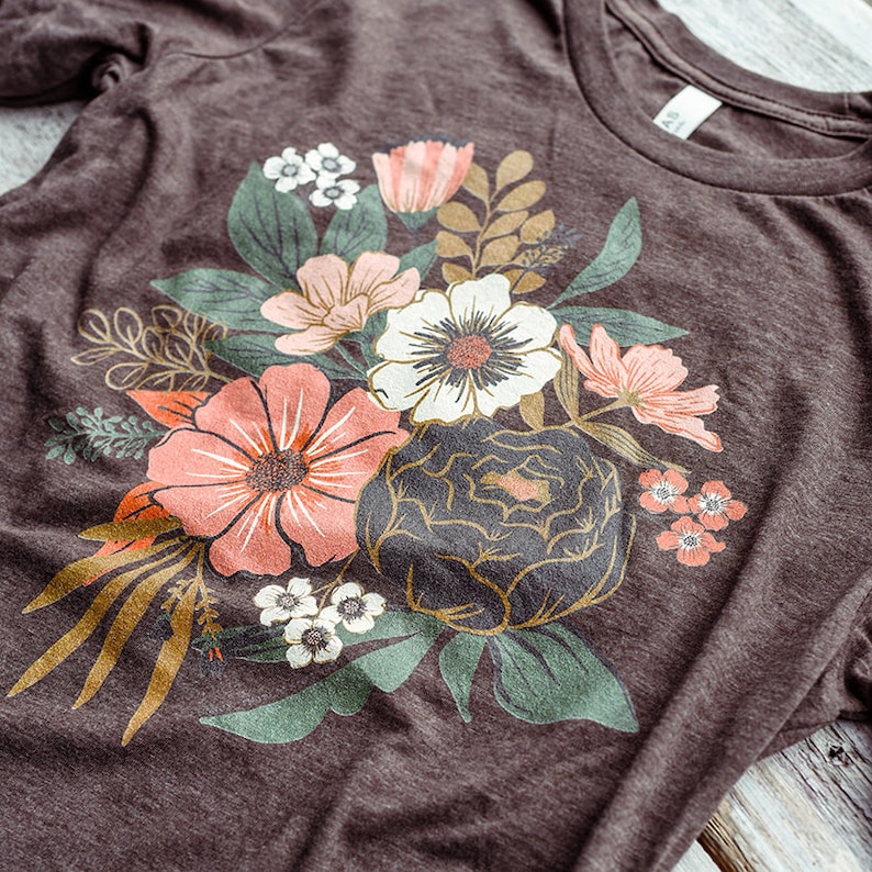 May include: A brown t-shirt with a floral print featuring a variety of flowers in shades of pink, white, and green. The flowers are arranged in a bouquet-like design.