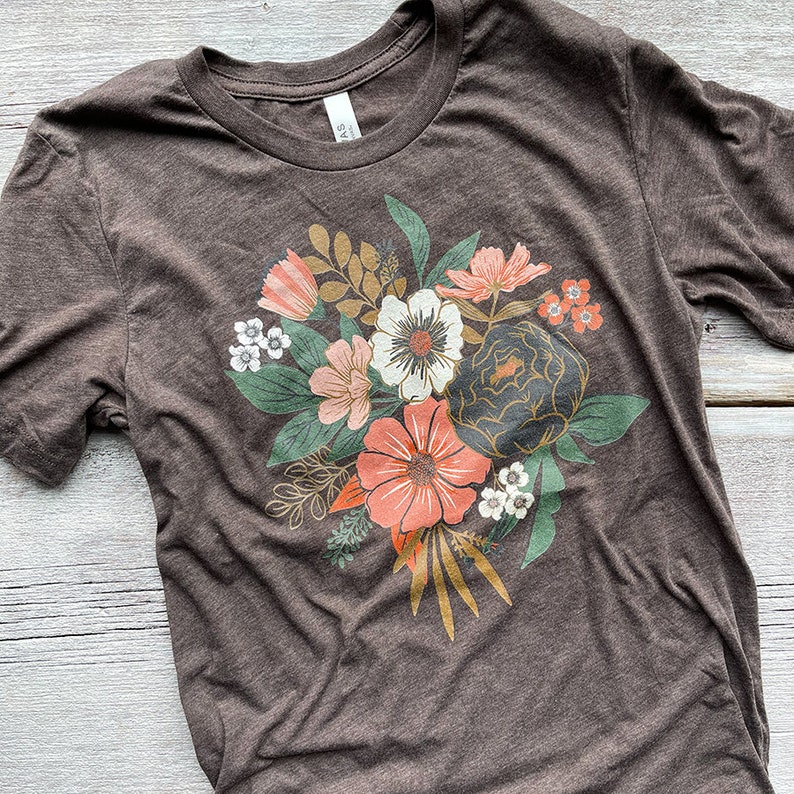 May include: A brown t-shirt with a floral print featuring a bouquet of flowers in shades of pink, orange, white, and green.