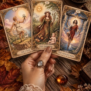 Puede incluir: Tres cartas de tarot tituladas The Fool, The Empress y The Hanged Man con ilustraciones detalladas de figuras en entornos naturales y arquitectónicos. Las cartas tienen bordes dorados ornamentados sobre un fondo de hojas de otoño.