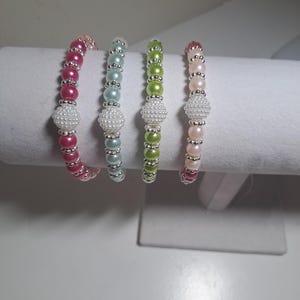 May include: Four beaded bracelets featuring alternating coloured pearl-like beads and small silver-toned metallic spacer beads. Each bracelet includes a larger, central white textured bead. The colours are hot pink, light blue, lime green, and pale pink.