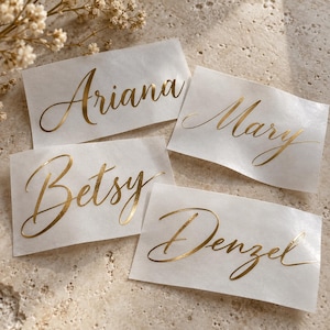 May include: Four rectangular white adhesive labels featuring the names Ariana, Mary, Betsy, and Denzel printed in a metallic gold cursive script font. The labels are arranged on a textured surface with small white flowers visible in the corner.