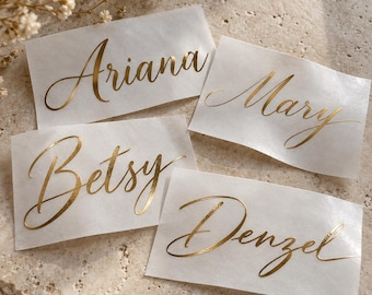 May include: Four rectangular white adhesive labels featuring the names Ariana, Mary, Betsy, and Denzel printed in a metallic gold cursive script font. The labels are arranged on a textured surface with small white flowers visible in the corner.