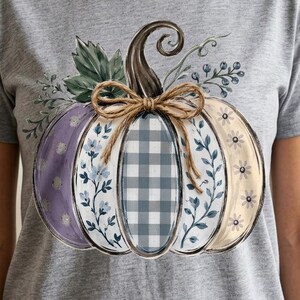 Puede incluir: Una camiseta de manga corta en color gris jaspeado con un gran gráfico de una calabaza. La calabaza está dividida en segmentos verticales con varios patrones, incluyendo morado, flores azules, cuadros vichy grises y blancos y crema.