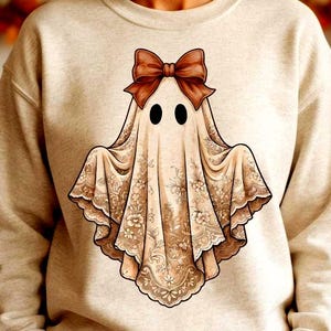 Puede incluir: Una sudadera de cuello redondo color beige con un gráfico central de un fantasma cubierto con una tela de encaje detallada en tono bronceado. El fantasma tiene dos ojos ovalados negros y un gran lazo marrón sólido sobre su cabeza.