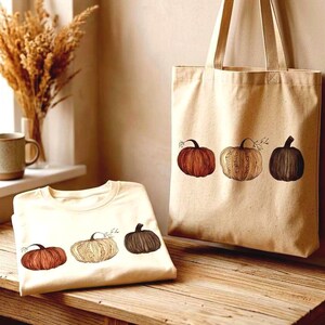 Puede incluir: Una bolsa de tela de algodón color crema y una camiseta plegada a juego, ambas con un estampado gráfico de tres calabazas en tonos naranja, beige y marrón oscuro. La bolsa cuelga de un gancho de madera y la camiseta descansa sobre una superficie.