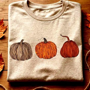 Puede incluir: Una camiseta de cuello redondo en color beige claro con un estampado gráfico de tres calabazas dispuestas en fila horizontal. La primera calabaza está delineada en marrón, la segunda es naranja con líneas detalladas y la tercera es rojo-naranja.
