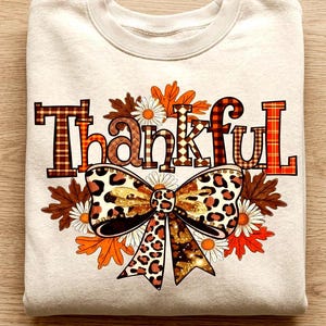 Puede incluir: Una sudadera de cuello redondo en color beige con la palabra Thankful en patrones de cuadros. Debajo del texto hay un lazo grande con estampado de leopardo rodeado de hojas de otoño naranjas y marrones y margaritas blancas en el frente.