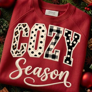 Puede incluir: Una sudadera roja de cuello redondo con la palabra COZY en letras grandes con varios patrones, incluyendo estampado de leopardo, lunares negros y cuadros escoceses en blanco y negro. La palabra Season está bordada debajo en letra cursiva blanca.