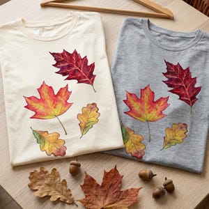 Puede incluir: Dos camisetas de cuello redondo, una color crema y otra gris jaspeado, cada una con un estampado estilo acuarela de cuatro hojas de otoño en tonos rojos, naranjas y amarillos. Las camisetas se muestran con una percha de madera y bellotas.