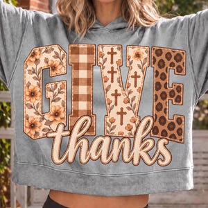 Puede incluir: Una sudadera con capucha gris que presenta el texto GIVE en letras grandes con patrones variados que incluyen flores, cuadros, cruces y estampado de leopardo, y la palabra thanks en cursiva debajo.