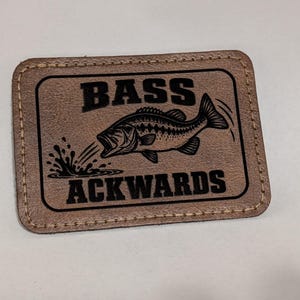 May include: A rectangular tan leather patch with rounded corners and beige stitching along the border. The patch features a black graphic of a jumping bass fish with water splashes and the black text BASS ACKWARDS printed in a bold, block font.