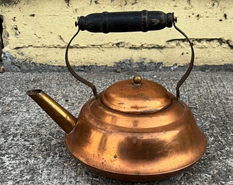 May include: A copper tea kettle with a wide, rounded base, a short angled spout, and a removable lid featuring a small brass knob. The kettle has a curved metal handle topped with a cylindrical, dark-stained wooden grip showing signs of wear.