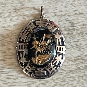 Puede incluir: Pendiente de plata con un diseño de esmalte negro y dorado de una persona tirando de un rickshaw. El pendiente está rodeado por un círculo de caracteres chinos.