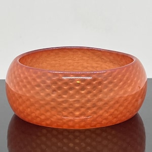 May include: A translucent orange bangle bracelet featuring a repeating textured diamond pattern across its entire outer surface. The circular accessory has a smooth, rounded profile and a glossy finish that catches the light, creating depth.