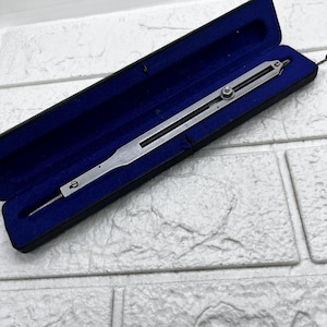 Puede incluir: Un compás de dibujo plateado vintage, abierto en su estuche negro con interior de terciopelo azul. La herramienta tiene una rueda de ajuste central y una punta afilada. El estuche es rectangular y tiene una tapa con bisagras.