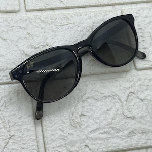 Puede incluir: Gafas de sol negras con lentes oscuras. Las gafas tienen una forma de montura redondeada y un diseño sutil de ojo de gato. Las patillas son finas y las lentes son oscuras, lo que proporciona un aspecto elegante y moderno. Las gafas están sobre un fondo de ladrillos blancos.