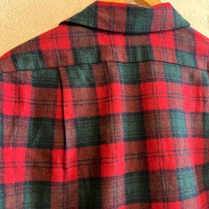 May include: A long-sleeved shirt featuring a red and dark green plaid pattern. The garment has a pointed collar, a button-down front, and a vertical pleat on the back yoke. The fabric has a soft, textured appearance consistent with flannel material.