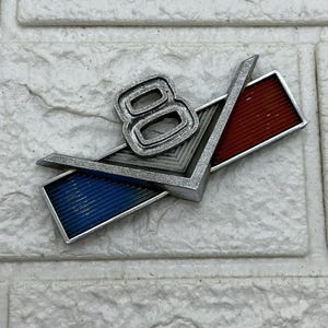 May include: A classic chrome emblem with the number "8" and a "V" design, incorporating red and blue sections. This metal emblem is likely for a vehicle.