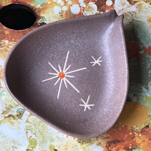 May include: A teardrop-shaped ceramic dish in a muted mauve tone with a textured, speckled finish. The center features a white starburst design with a small orange circular accent, accompanied by two smaller white star motifs to the right side.