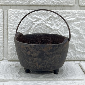 May include: A small, dark brown cast iron cauldron with a rough, textured surface. The pot features three short, sturdy legs at the base and a thin, arched metal handle attached to the sides. The interior is deep and dark with a weathered appearance.