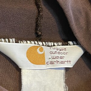 May include: Close-up of a brown garment with a tag. The tag features a gold logo and the words "rugged outdoor wear carhartt" in a dark font. The garment appears to be made of a soft, durable material.