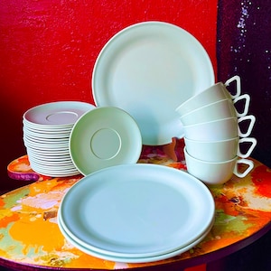 May include: A set of pale green dinnerware including a large round serving platter, two dinner plates, a stack of small saucers, one individual saucer, and a vertical stack of four cups with distinct triangular handles and smooth surfaces.