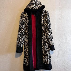 May include: A long, hooded coat with a zebra print pattern in black and white. The coat has black trim and a red interior lining. The coat is hanging against a neutral-colored wall.