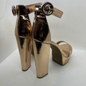May include: Rose gold platform heels with a thick block heel and ankle strap. The shiny metallic finish reflects light, and the buckle adds a touch of detail. The open-toe design and platform sole provide height and style.