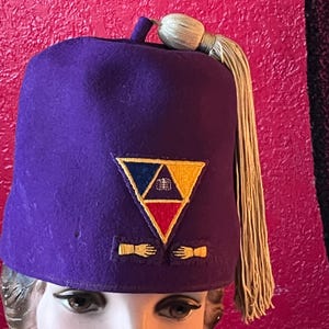 May include: A purple fez hat with a triangular emblem featuring red, yellow, and blue sections. The emblem includes a symbol and two gold-colored hand designs. A gold tassel hangs from the top of the hat, which is displayed on a mannequin head.