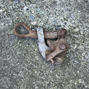May include: A weathered metal clamp, featuring a loop and a hinged section. The clamp, showing signs of rust, is set against a textured grey surface, suggesting outdoor use.