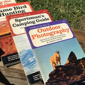 May include: A stack of vintage outdoor skill books with colorful covers. Titles include "Game Bird Hunting," "Sportsman's Camping Guide," and "Outdoor Photography." The "Outdoor Photography" book features a mountain goat on the cover.