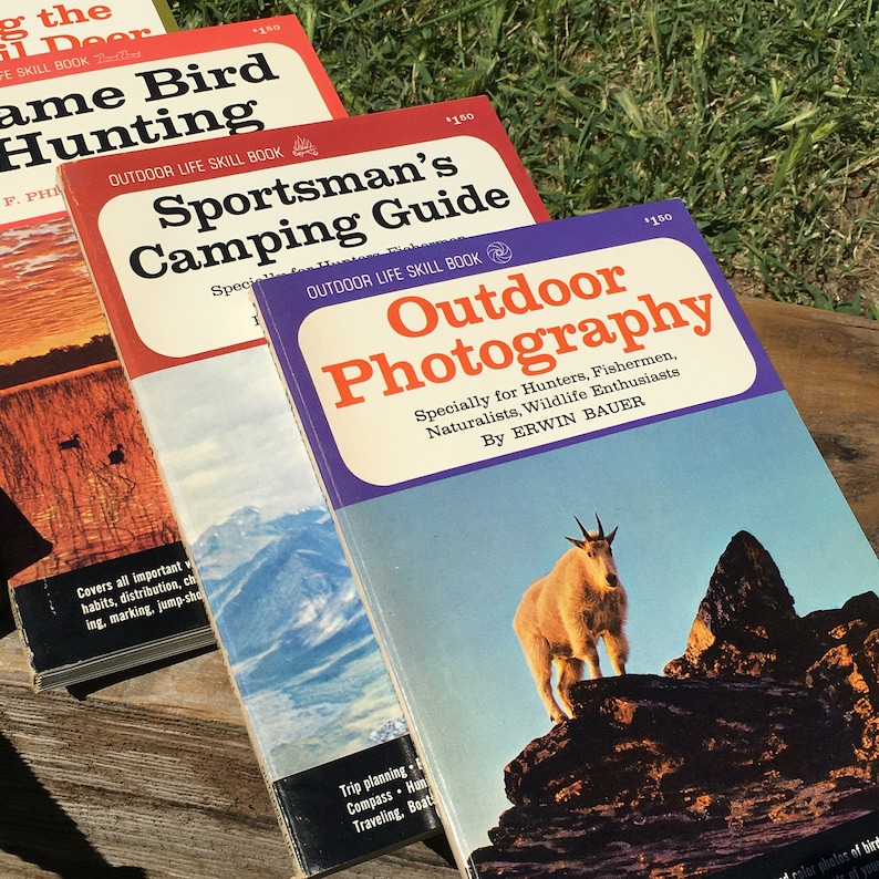 May include: A stack of vintage outdoor skill books with colorful covers. Titles include "Game Bird Hunting," "Sportsman's Camping Guide," and "Outdoor Photography." The "Outdoor Photography" book features a mountain goat on the cover.