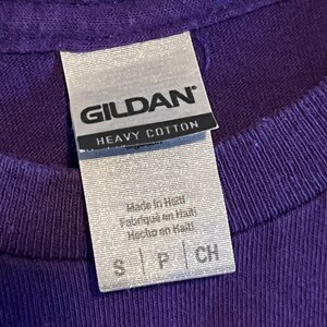 May include: A white tag with black text that reads "GILDAN HEAVY COTTON" and "Made in Haiti" in smaller text. The tag is attached to a purple t-shirt.