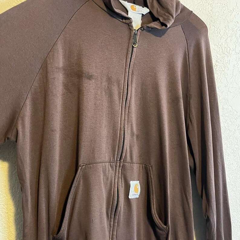May include: A brown zip-up hoodie with a hood and a kangaroo pocket. The hoodie has a full zipper closure and a small logo patch on the pocket. The fabric appears to be a soft, knit material. The hoodie is hanging against a neutral background.