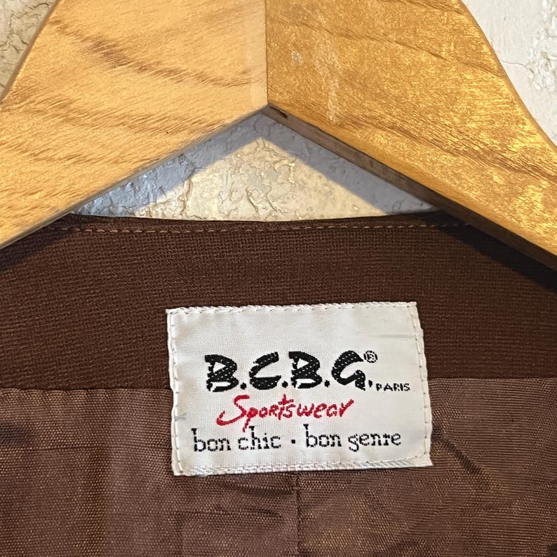 Puede incluir: Una prenda marr&oacute;n con una etiqueta rectangular blanca con texto en negro y rojo que dice B.C.B.G. PARIS, Sportswear y bon chic bon genre. La etiqueta est&aacute; cosida sobre la tela interior, la cual tiene una textura tejida visible.