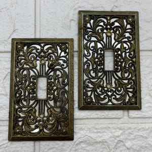 Puede incluir: Dos cubiertas de interruptores de luz antiguas de color latón. Cada cubierta presenta un diseño calado ornamentado de hojas arremolinadas y motivos florales. Las cubiertas rectangulares tienen una abertura central para el interruptor y están montadas en una pared de ladrillos blancos.