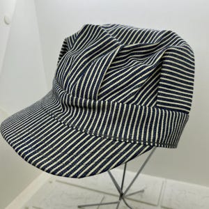 May include: A blue and white striped cap with a short brim. The cap has a rounded crown and is made of a fabric with a repeating stripe pattern. The cap is displayed on a metal stand.