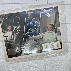 May include: A vintage lobby card for the film Moon Zero Two featuring three individuals in white and blue space suits inside a spacecraft. The text includes the film title Moon Zero Two and the names James Olson, Catherina von Schell, and others.