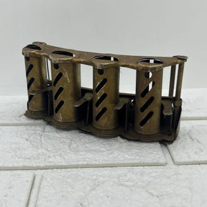 May include: A vintage brass coin dispenser featuring four cylindrical slots. Each slot has diagonal cutouts and a spring-loaded mechanism. The dispenser is designed to hold and dispense coins, possibly for a cash register.