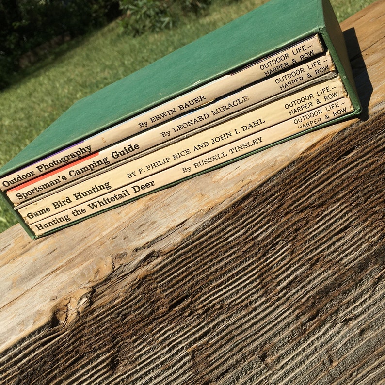 May include: A collection of vintage outdoor life books with green covers and titles such as "Outdoor Photography" and "Sportsman's Camping Guide." The books are stacked on a weathered wooden surface, with the titles and authors visible.