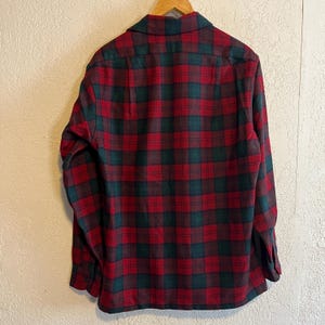 May include: A long-sleeved button-down shirt featuring a classic plaid pattern in deep red and dark green. The shirt has a standard collar, a buttoned front placket, and buttoned cuffs, displayed hanging on a wooden hanger against a textured wall.